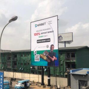 Portrait billboard advertising NIRIBET along Obafemi Awolowo Way, Ikeja, Lagos, featuring a high-traffic area with buses and pedestrians.