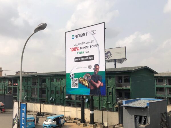 Portrait billboard advertising NIRIBET along Obafemi Awolowo Way, Ikeja, Lagos, featuring a high-traffic area with buses and pedestrians.