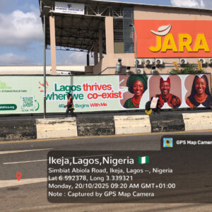 Wall-drape billboard on Simbiat Abiola Road, Ikeja showing a long horizontal ad reading “Lagos thrives where we co-exist” beside a retail storefront.